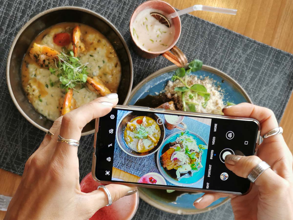 Mão segurando um celular e tirando foto de dois pratos de comida em um restaurante na Praia Brava, Itajaí.