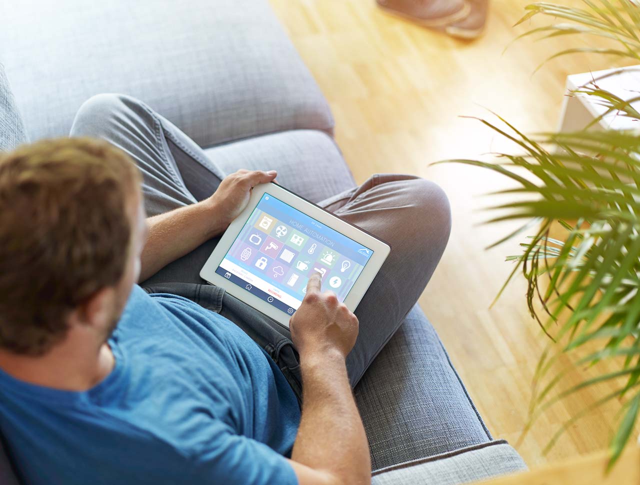 Foto de um homem sentando em um sofá mexendo no tablet como se estivesse configurando os sistemas de automação residencial.