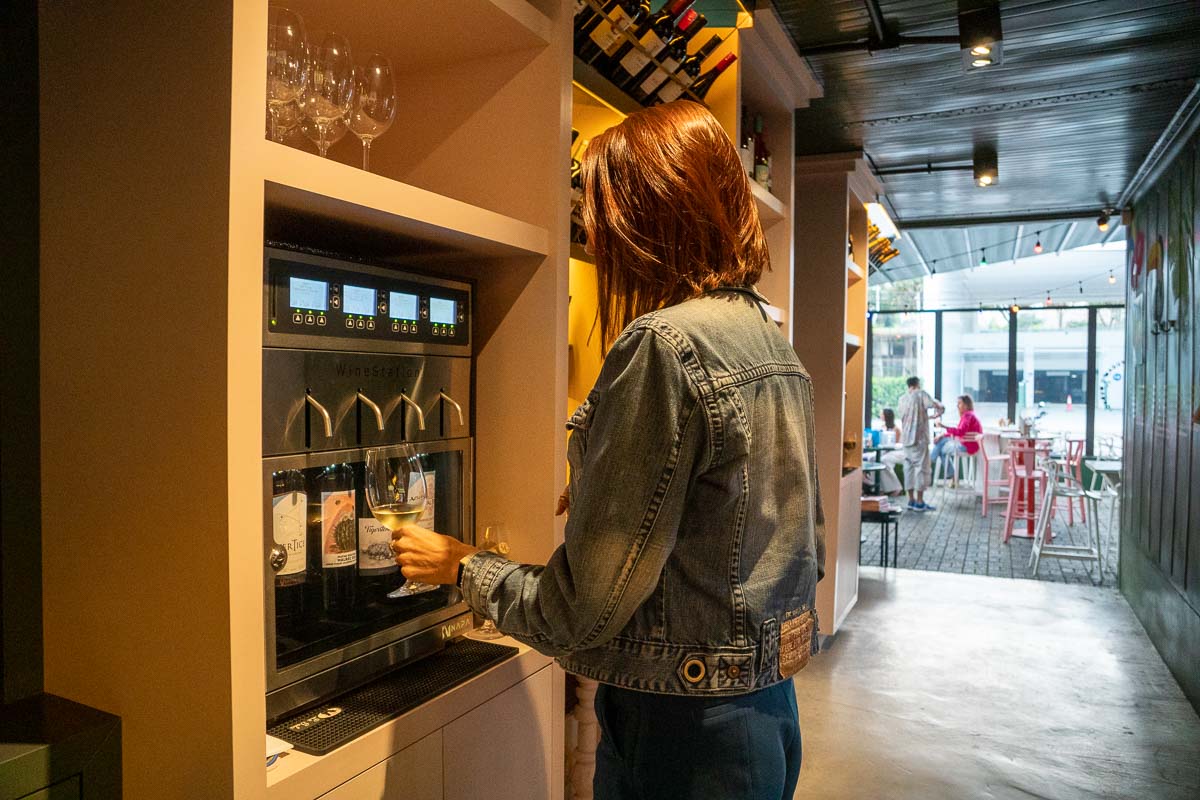 Mulher comprando vinho em uma máquina de self-service onde a dose do vizinho é dispensada automaticamente.